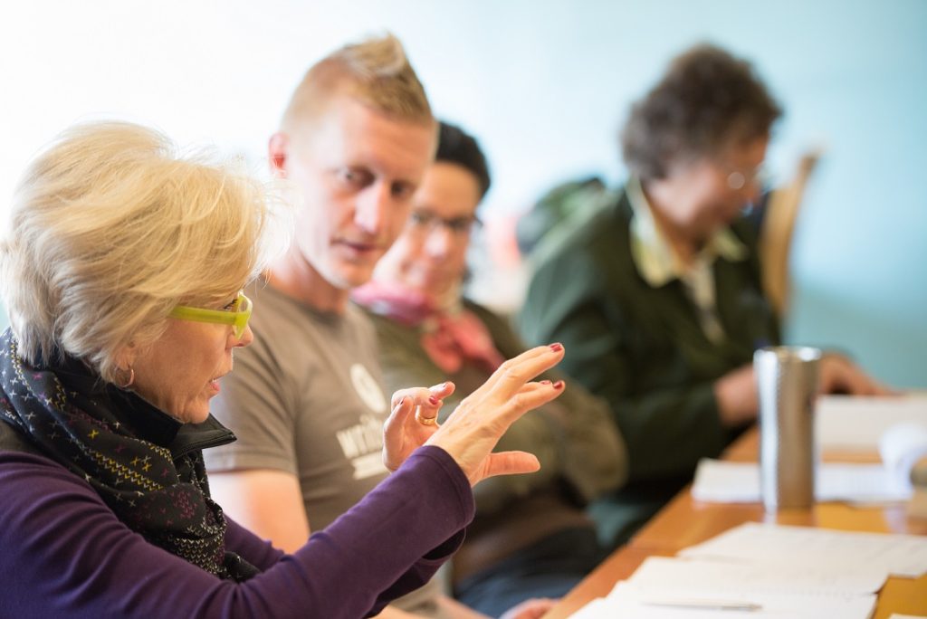 A woman with green glasses and a purple top holds up her hands to illustrate a point as she speaks to a room of people sitting around a table. Several people sit further away, looking at her or down at the table. Their faces are blurred slightly. 