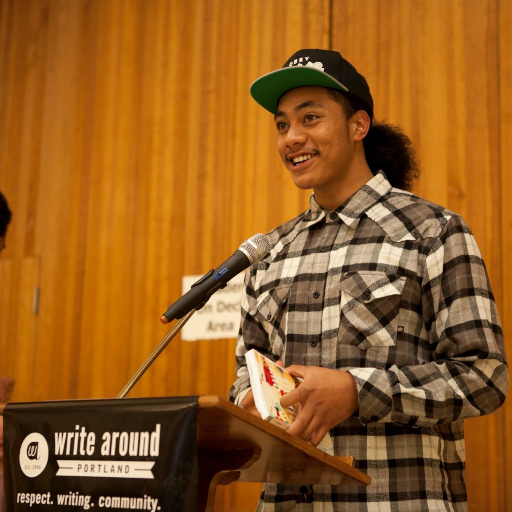 A young man with a baseball hat stands at a podium in front of a microphone, holding a book in his hands. He smiles. 