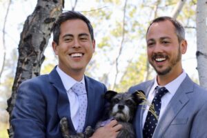Two smiling men in suits and ties snuggle a grey dog with white snout between them. 