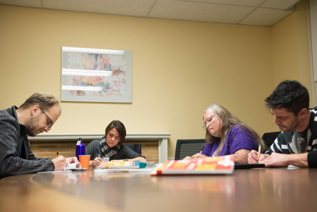 Four people sit around a table with pens in their hands, writing on journals. They appear to be very focused on their writing.