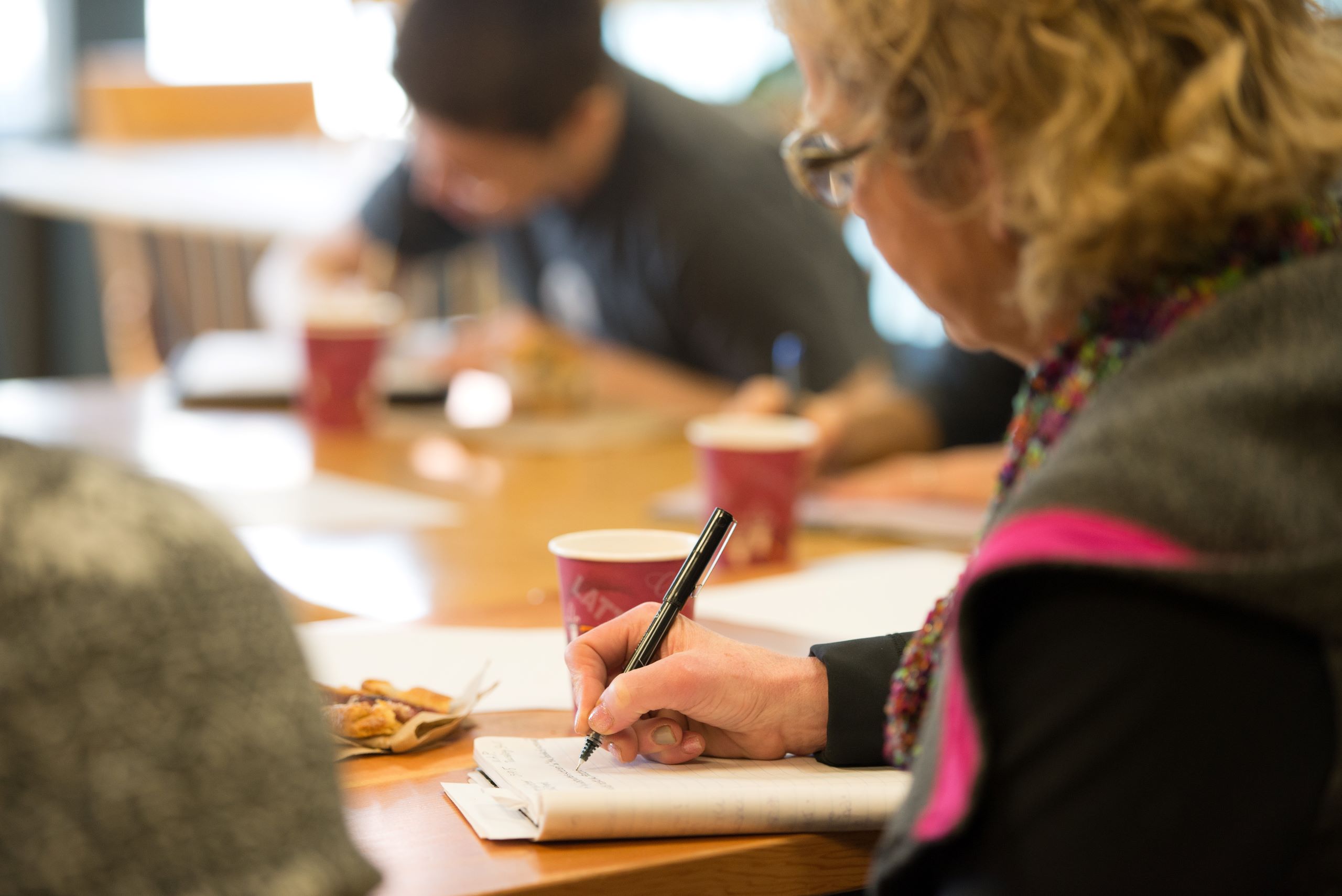 A woman holds a pen in her hand and sits at a table with other people writing in the background. Her face is obscured.