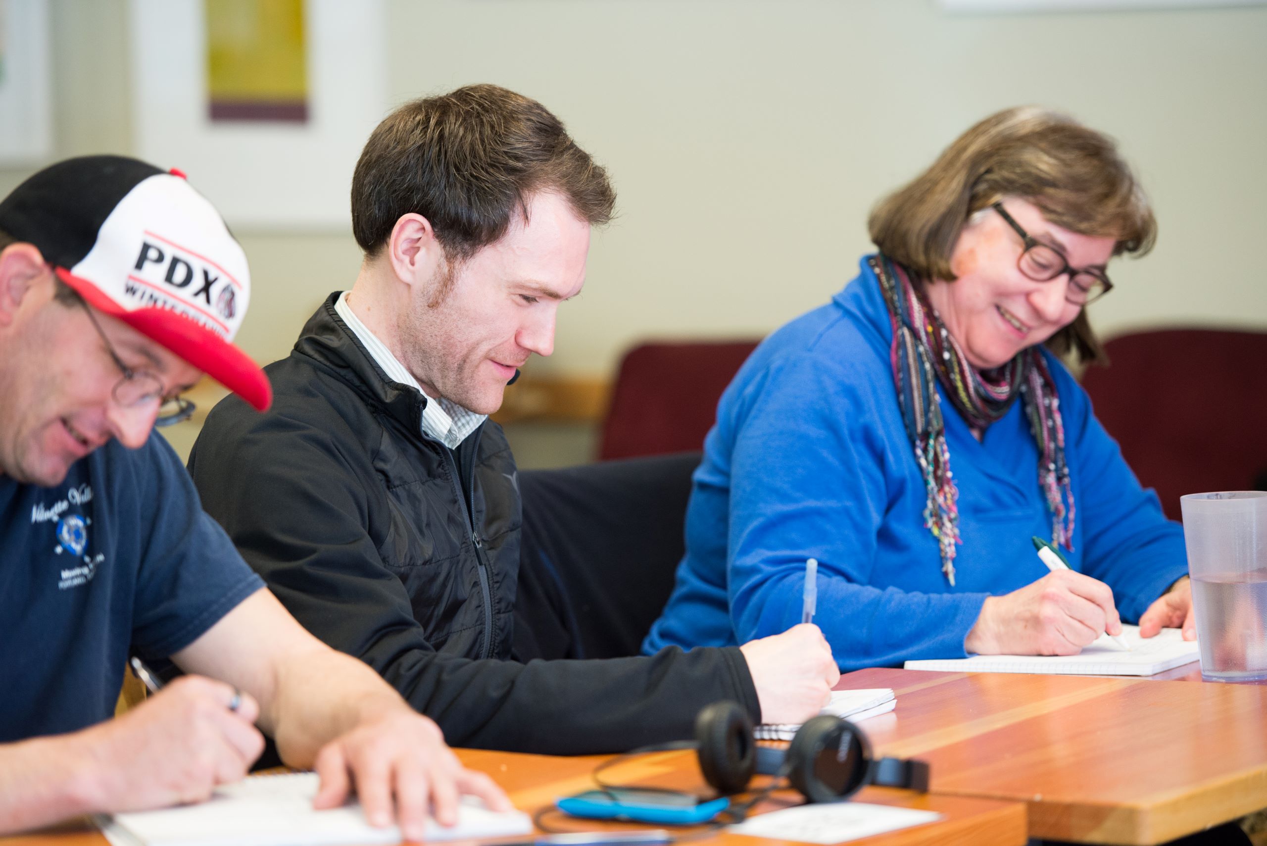 Three people sit at a table with pens in their hands, writing. All of them smile.