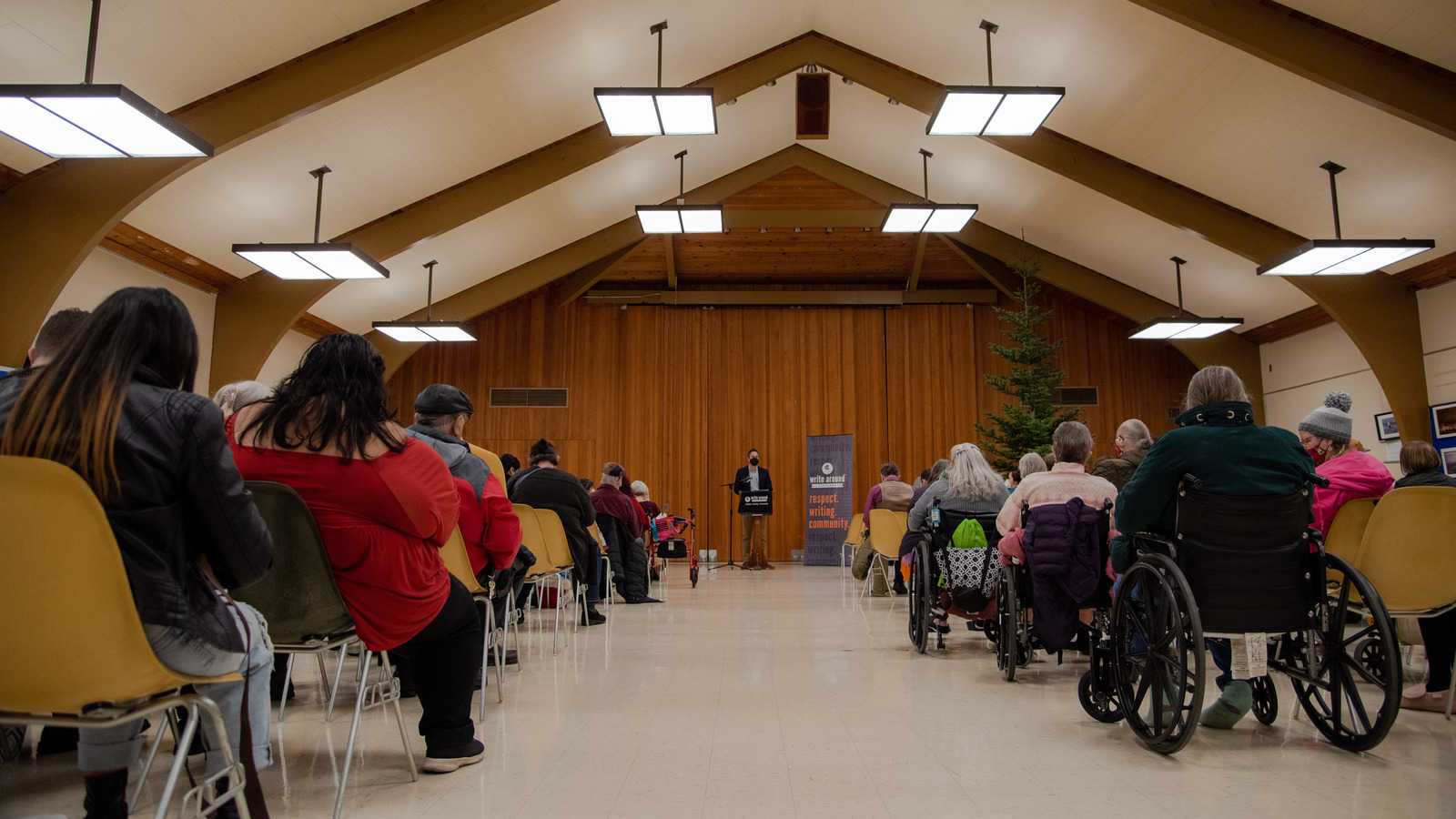 A man stands at a podium wearing a face mask. There's a sign next to him that has the Write Around Portland logo and the words respect writing community in orange. The photograph has been taken at a low angle facing upward down a long aisle between two sections of chairs. Some people sit use wheelchairs while others sit in yellow plastic chairs. The floor below them gleams under fluorescent lighting. 