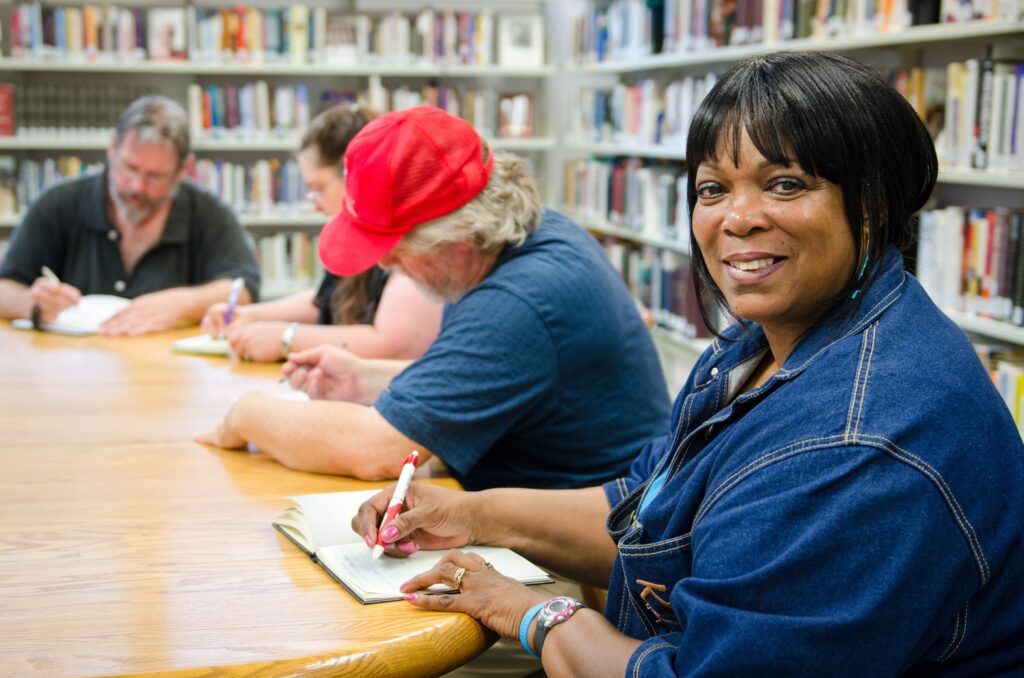 A woman smiles as she holds a pen with a journal underneath. In the background, three other people seem to be busy writing. The backdrop seems to be a library.