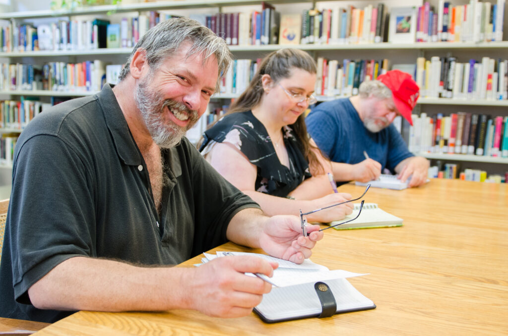 Three people sit at a table with journals in front of them. Two of them are writing. The third - a man closest to the camera - has taken off his glasses and smiles at the camera. 