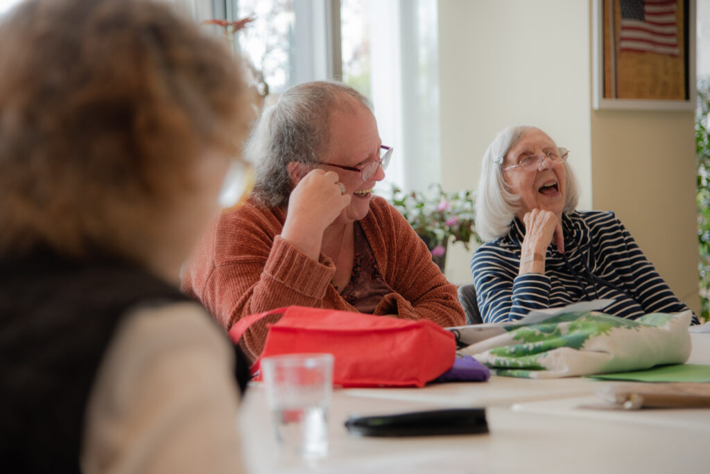 A group of three people (one obscured in the foreground) sit around a table. Two of the people are laughing. On the table, there are various tote bags, a water glass, and papers. 