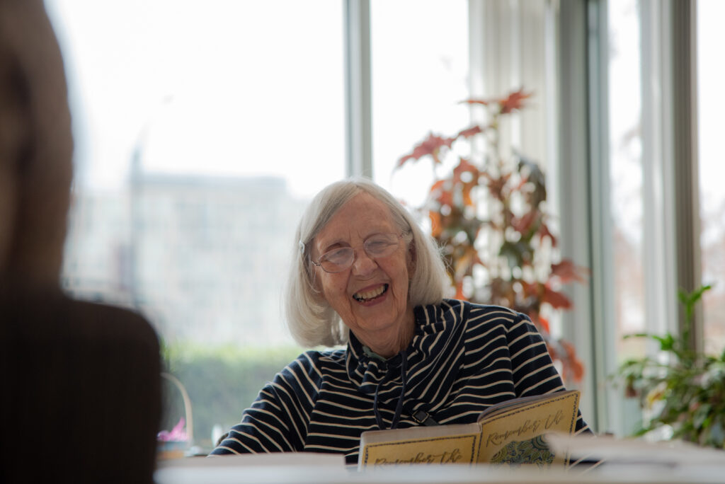 An older woman with white hair and glasses laughs while holding a journal. 