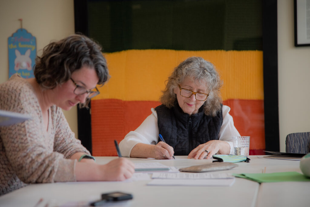 Two women sit at a table with pens, writing on pieces of paper. They appear to be concentrating. One of them smiles.