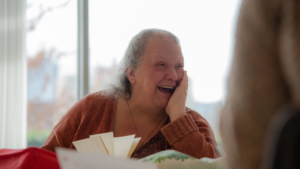 An older woman smiles with elation. An open journal is front of her and she has her hand on her cheek as if she can't stop laughing.