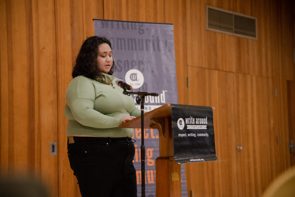 A woman of color stands in front of a podium, reading her work. 