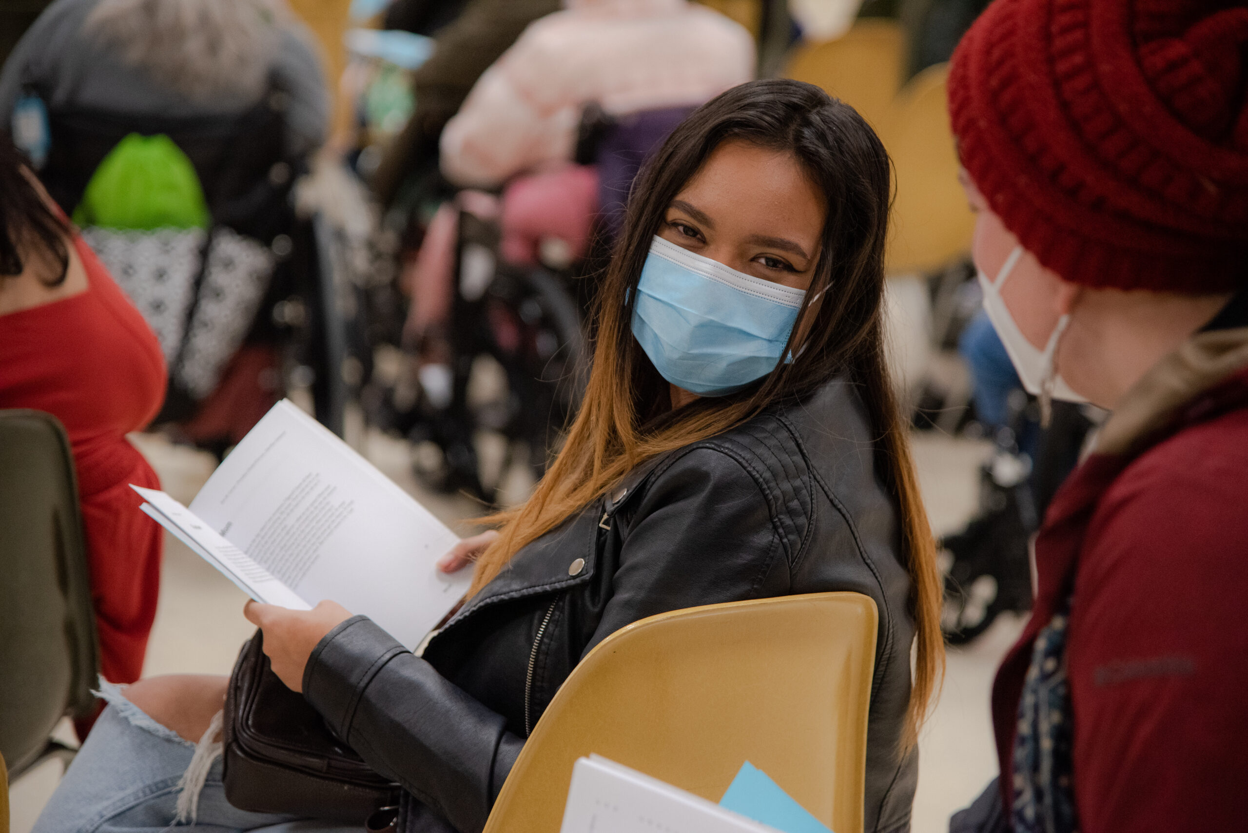 A woman with a blue facemask looks into the camera. Her eyes indicate that she's smiling. She is sitting in a chair with a Write Around Portland anthology open. There are other audience members sitting around her, their faces obscured. 