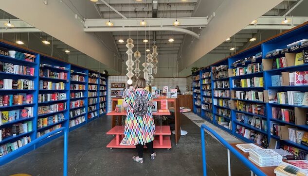 Photograph of a person from behind in a rainbow colored dress and backpack. They are standing at a pink bookshelf filled with books. The large, industrial space with high, white ceiling is lined on the left and right with bright blue bookshelves, full of colorful books. Paper sculptures made from book pages hangs from the ceiling.