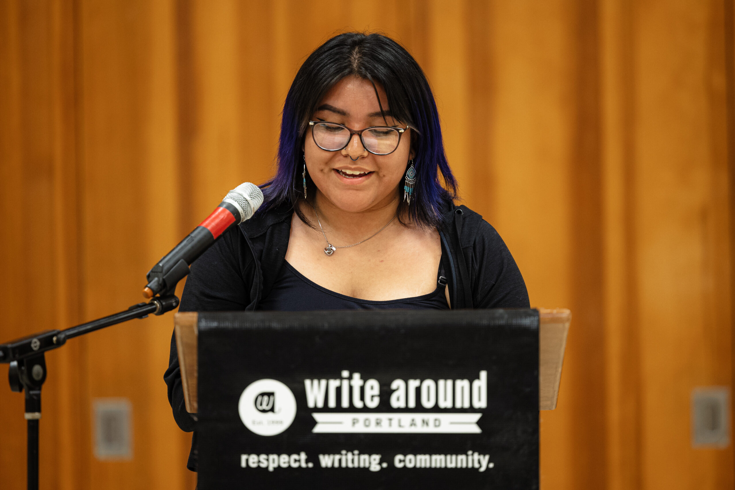 A woman stands in front of a microphone and smiles. She is looking down and seems about to begin to read. She has purple streaks in her black hair, wears glasses, and has turquoise earrings on.