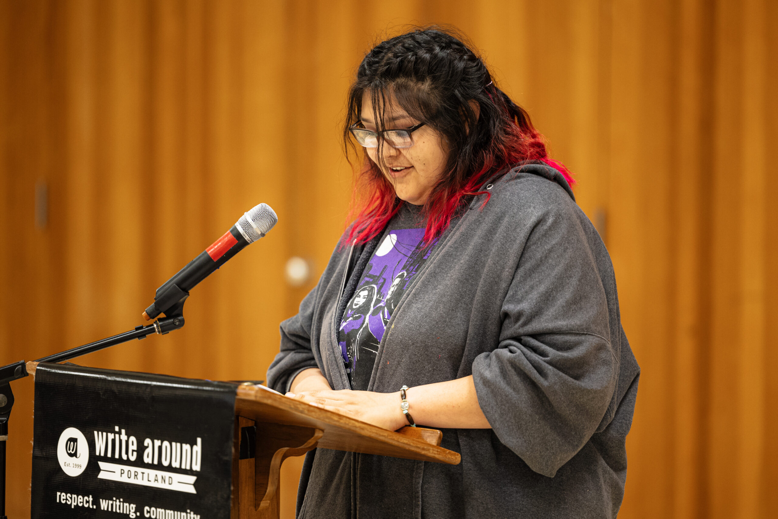 A woman of color in glasses stands in front of a microphone looking down. She smiles faintly.