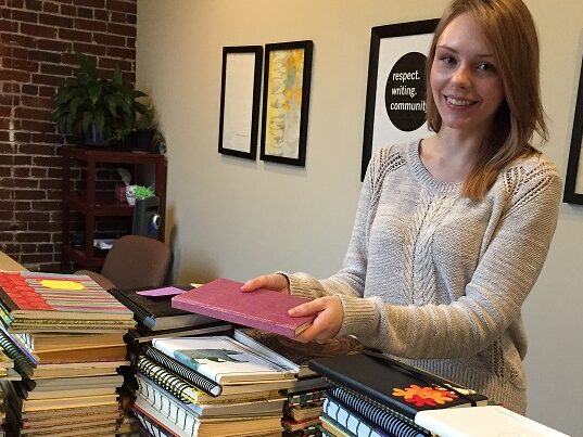 Person with dark blond shoulder-length hair and tan sweater smiles at the camera and places a journal on top of a stack of other journals.