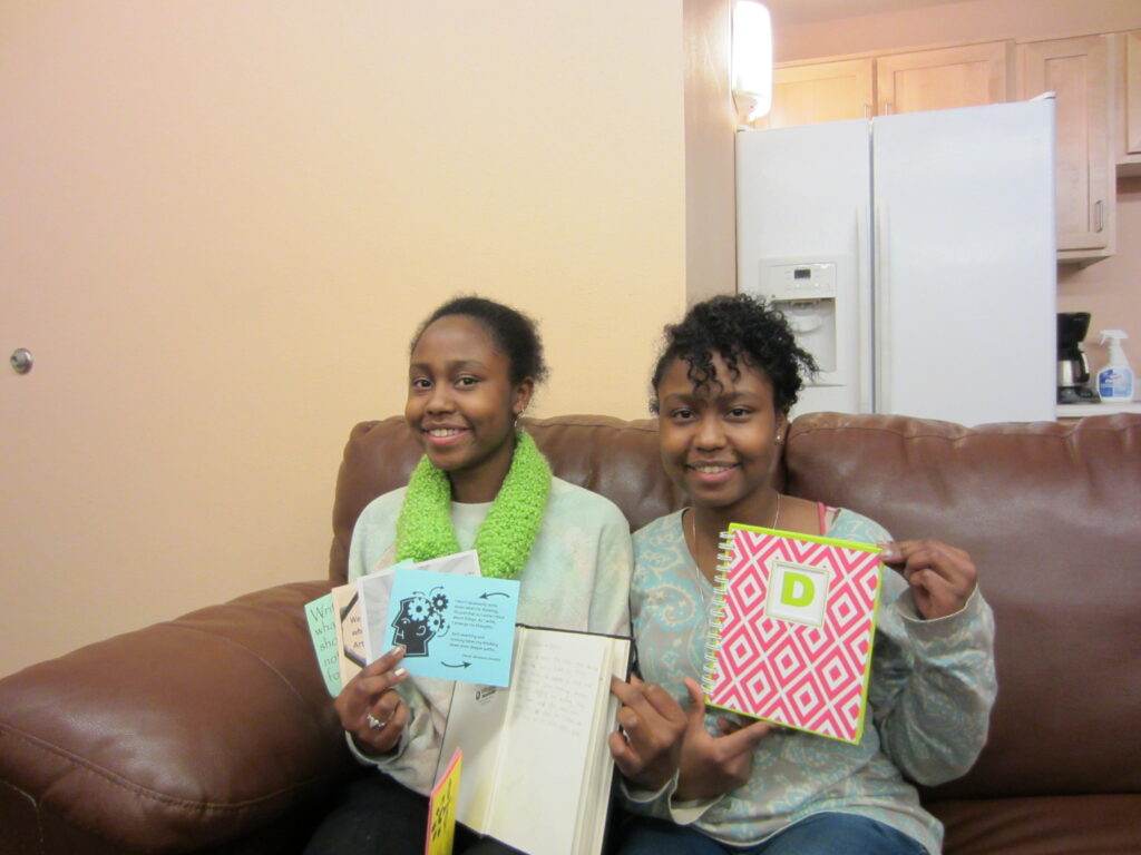 Two Black teenaged girls sit on a leather sofa and hold up postcards and journals. They smile at the camera.