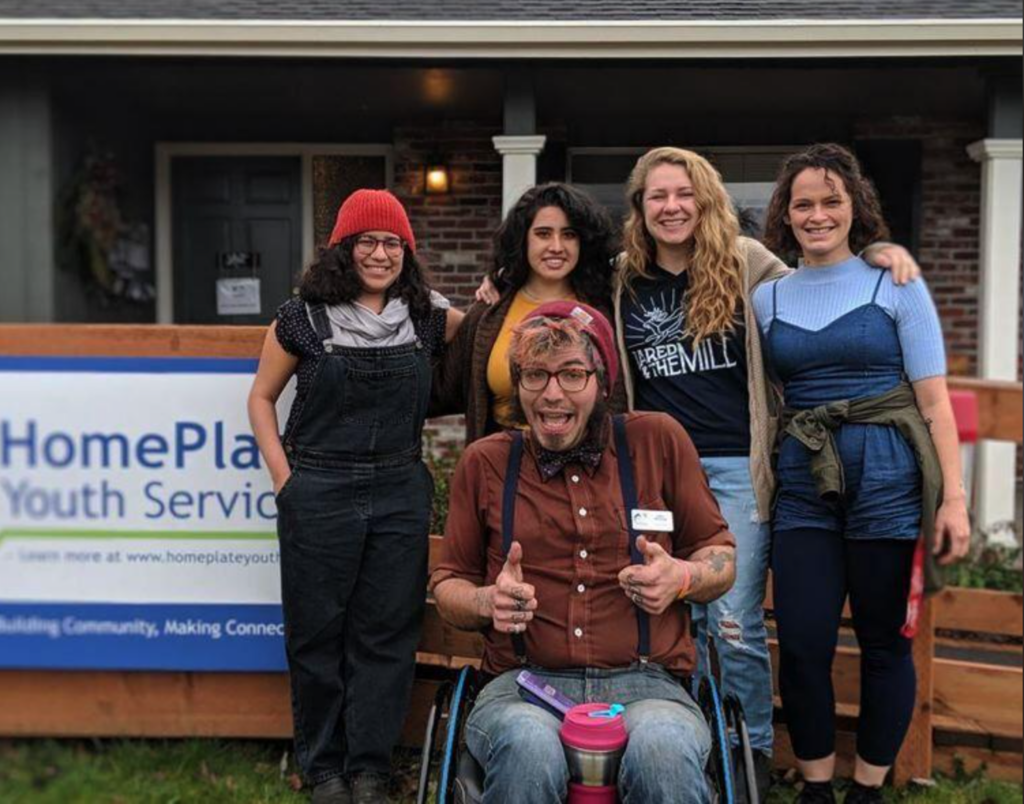 A group of five people in front of a brick house smile at the camera. Four are standing in a row, and one is in a wheelchair in front of them, giving two thumbs up. Next to them is a HomePlate Youth Services banner.