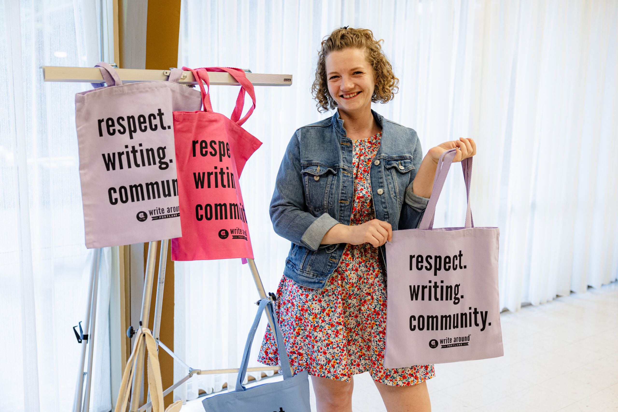 A woman stands in front of a curtained window and holds a lavender totebag with the words "respect. writing. community" emblazoned on it in bold black font. Two other bags are hanging next to her.