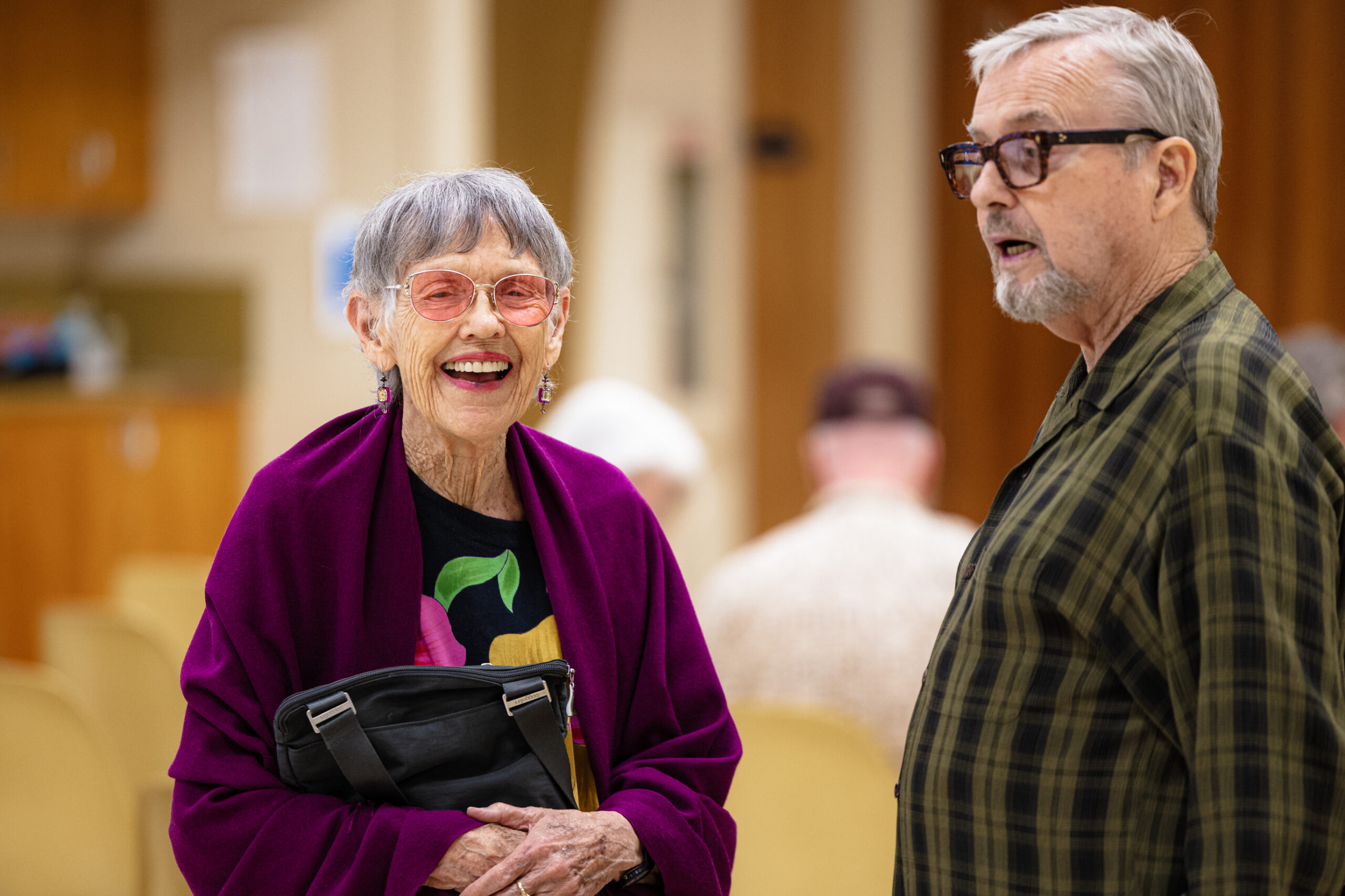 An older woman with pink-tinted eyeglasses smiles into the camera. She has purple earrings and a purple shawl on. In the foreground stands an older man with tortoise eyeglasses; his face is partially obscured.