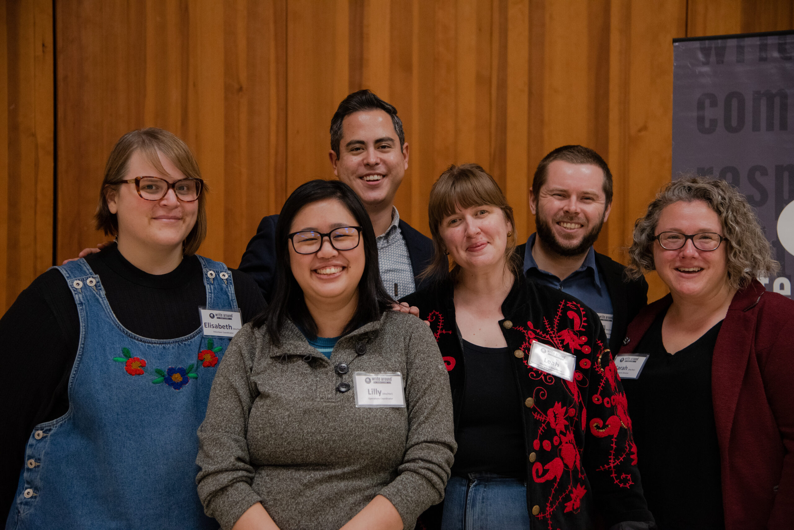 A group of Write Around Portland staff members pose together in front of a honey-colored wooden wall.