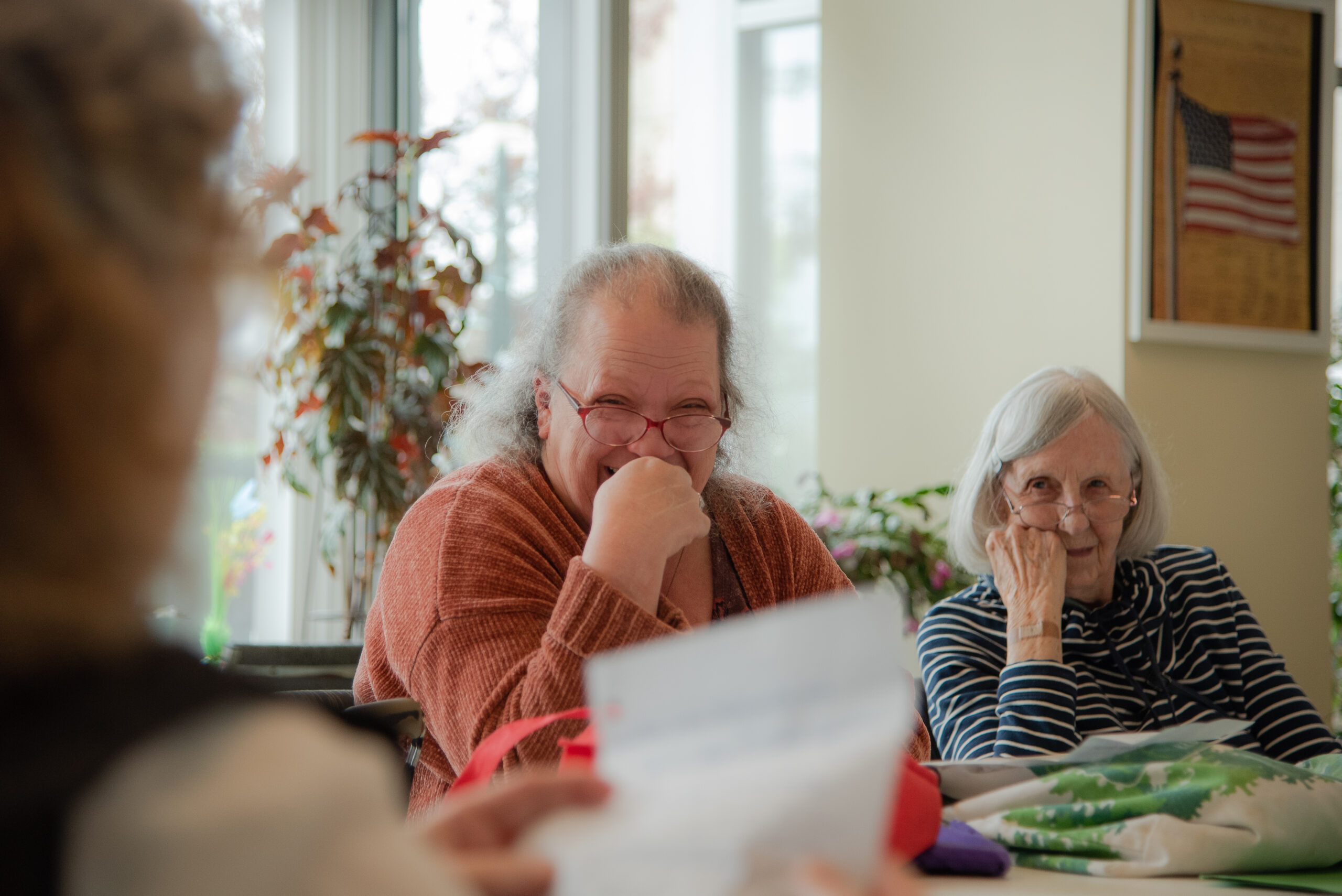 A person in an orange sweater puts their hand to their mouth and giggles as someone off-screen holds up a piece of paper, reading their work. A white-haired woman with glasses sits to the person's left and smiles faintly, her head resting on her knuckles. 