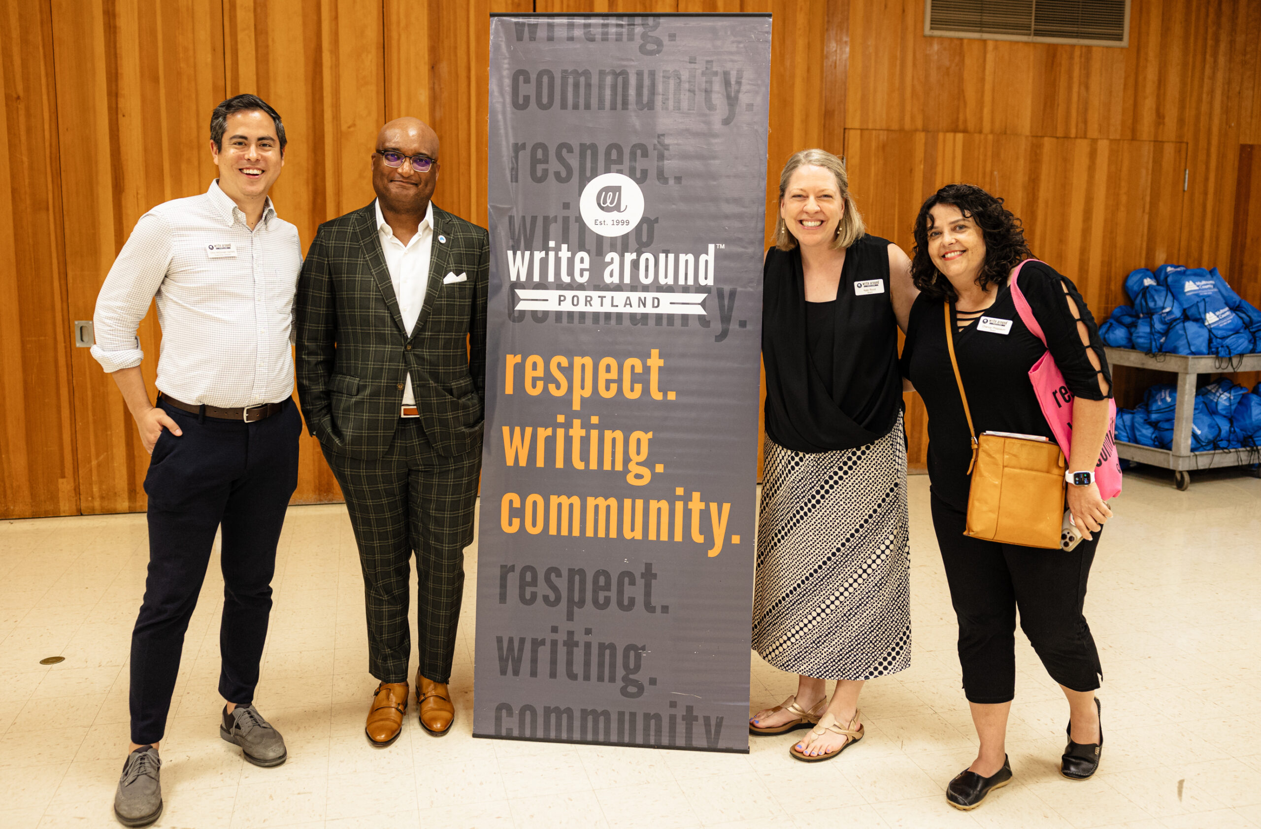 Four people smile and stand around a gray banner that says "respect. writing. community." in orange. 