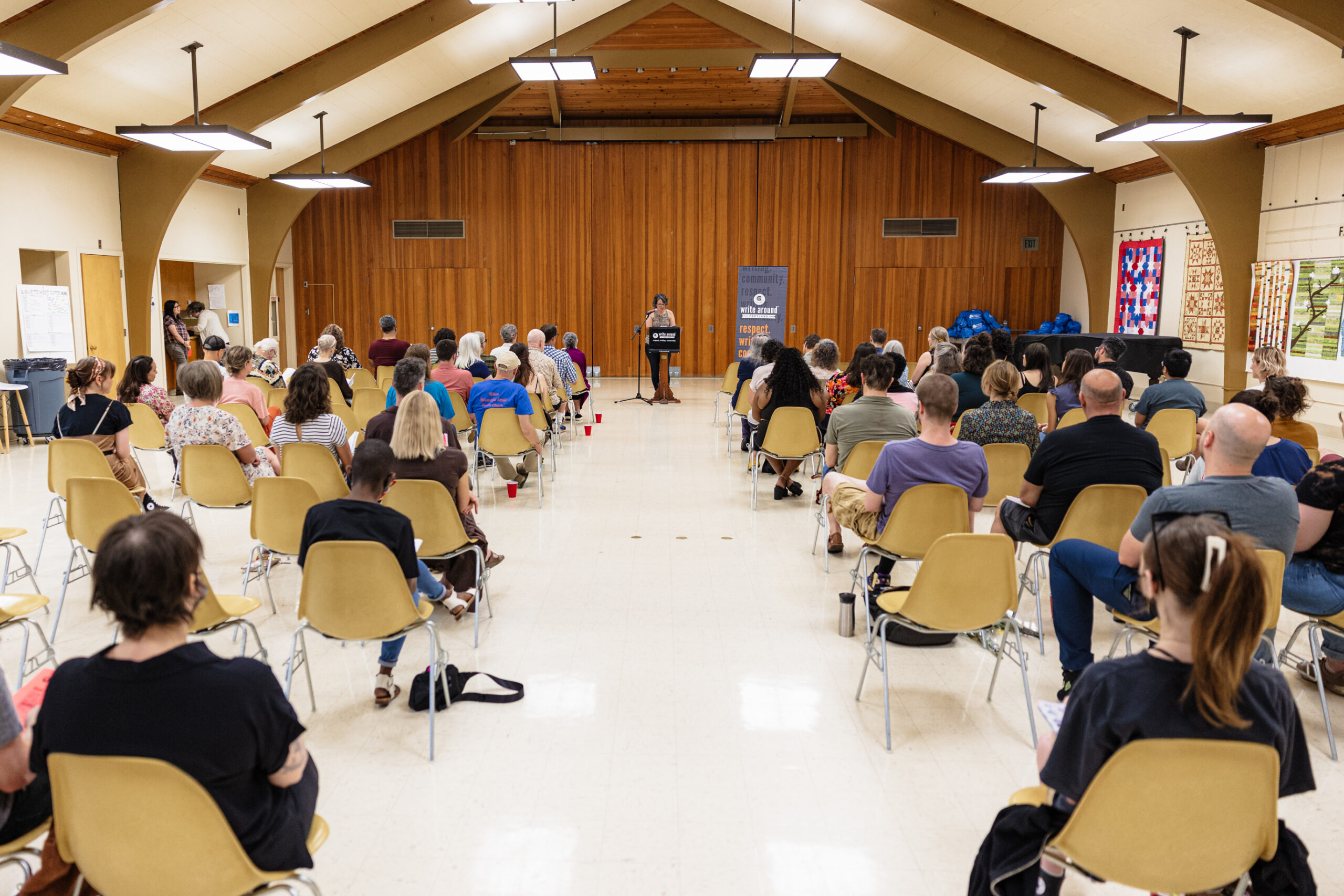 A woman stands in front of an audience at a podium with a black banner with the Write Around Portland logo on it. There is a microphone next to her. The room looks full with a wide aisle down the middle. The room is lit by fluorescent lighting and there is a wooden wall behind her.