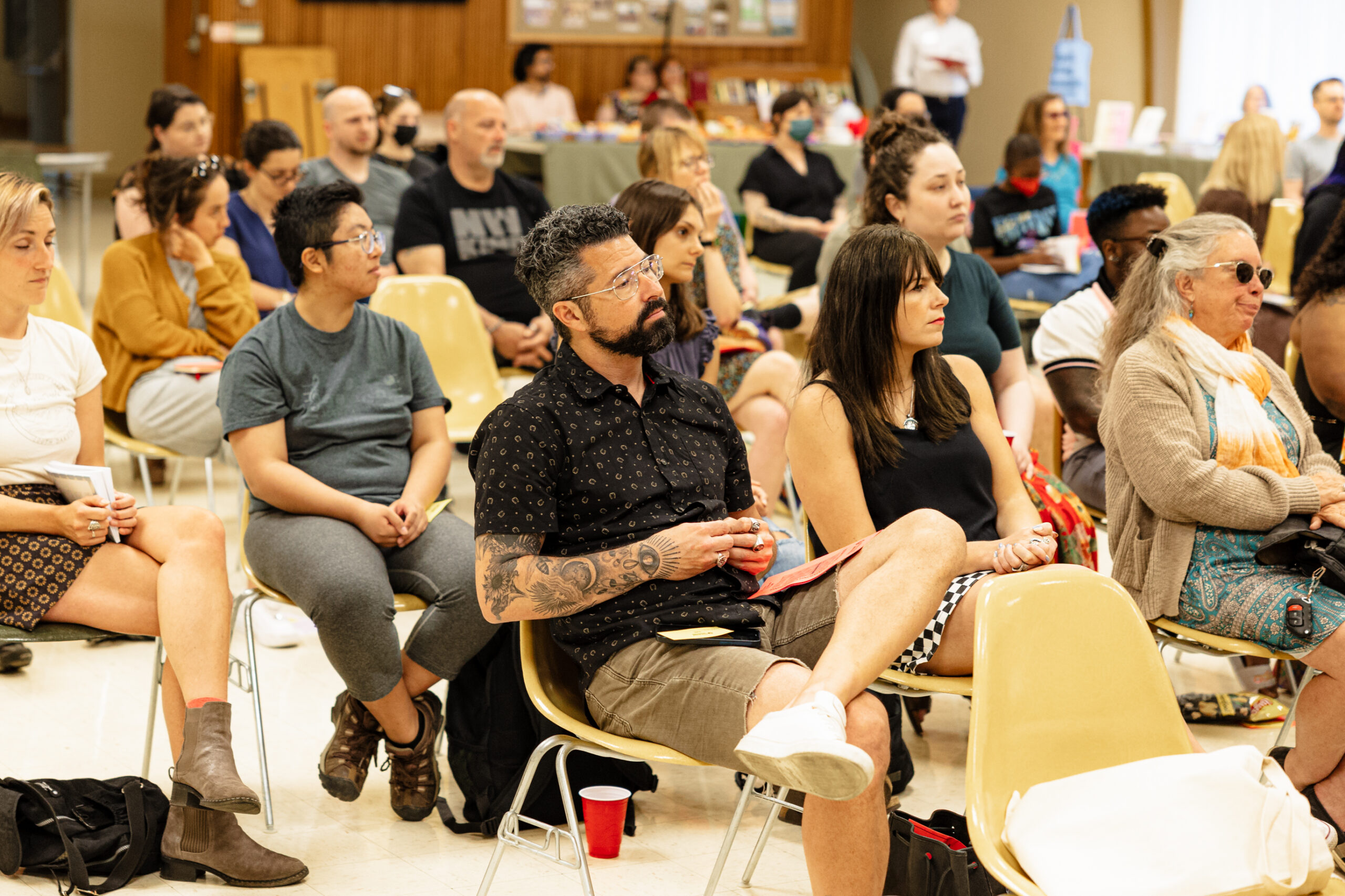 An audience of many people sit in yellow plastic chairs. Expressions on their faces range from serious to contemplative. 