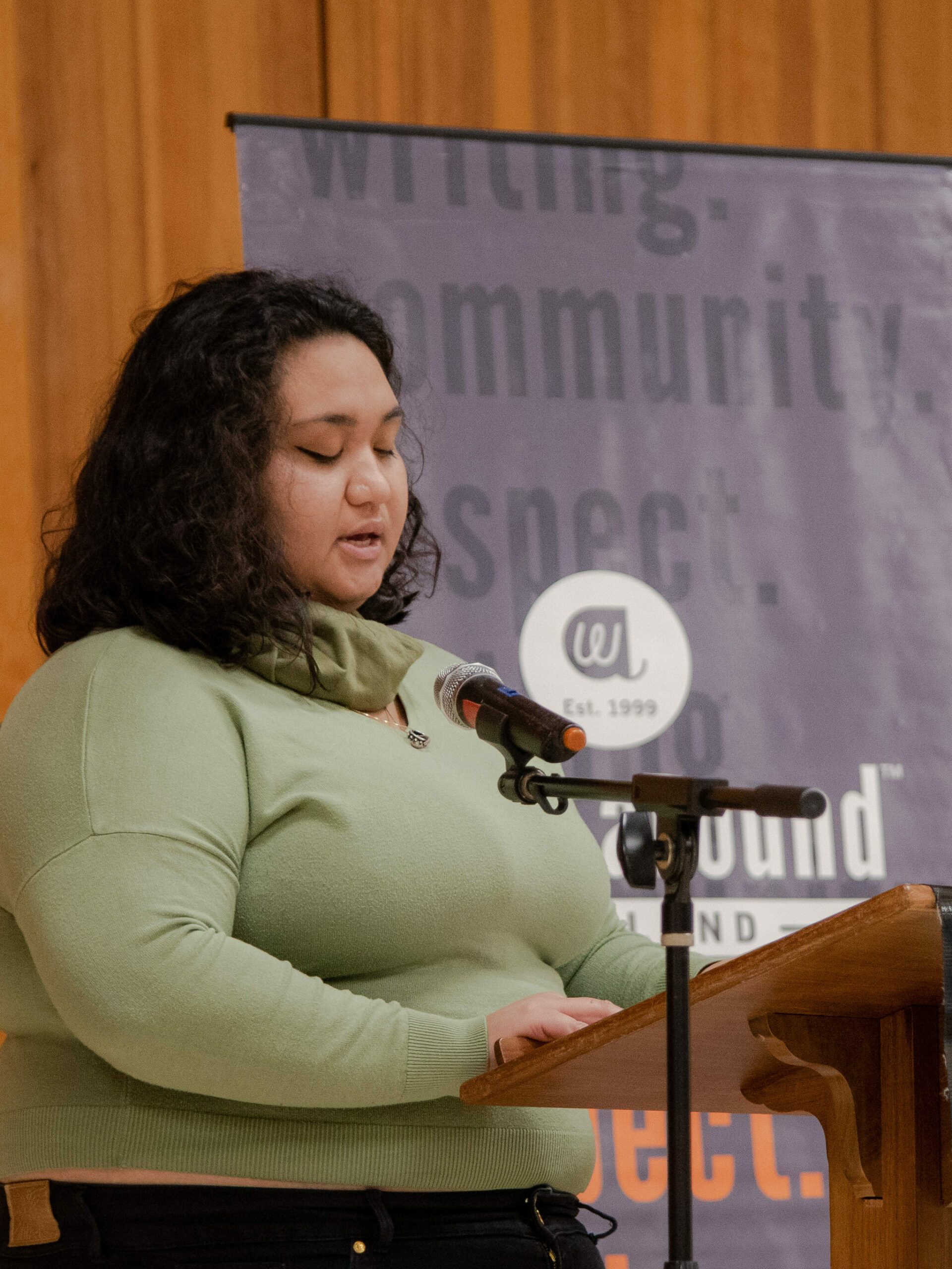 A woman in a sage green shirt reads from her published piece at a microphone.