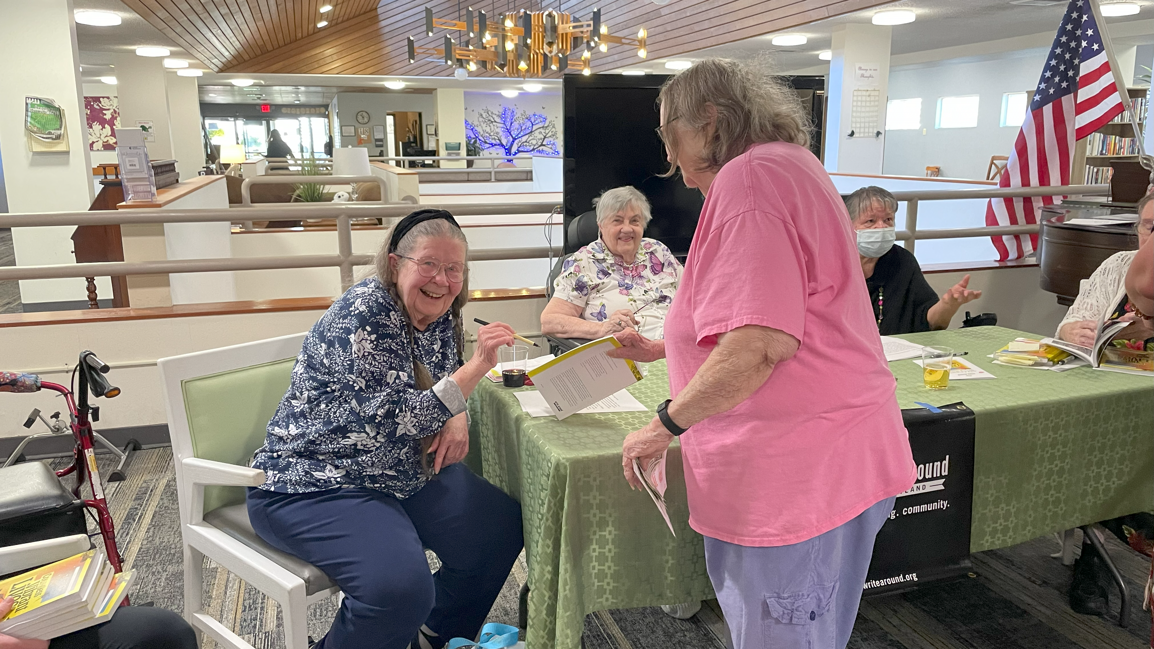 Several ladies sit at a Write Around Portland table smiling in their assisted living community. One holds a pen and is about to autograph a book.